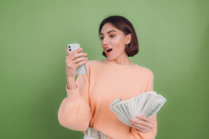 Young woman in casual peach sweater  isolated on green olive background excited holding black screen phone and money amazed surprised winner  emotions